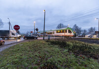 Widok z ziemi na przejazd kolejowo-drogowy, oczekujące na przejazd samochody i przejeżdżający po torach pociąg osobowy. 