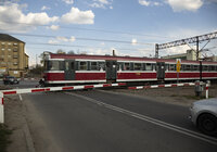 Widok z ziemi na przejazd kolejowo-drogowy, oczekujący na przejazd samochód i przejeżdżający po torach pociąg osobowy. 