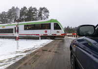 Widok z ziemi na przejazd kolejowo-drogowy, oczekujące na przejazd samochody i przejeżdżający po torach pociąg osobowy. 